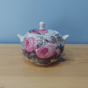 Puede incluir: Un azucarero de cerámica blanca con tapa, con un diseño floral de rosas rosas y hojas verdes.