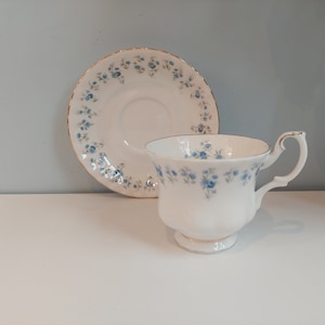 May include: A white teacup and saucer with a delicate blue floral pattern. The cup has a rounded shape and a handle, while the saucer has a scalloped edge.