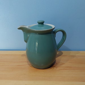 May include: A teal ceramic creamer with a white rim and lid. The creamer has a rounded body and a handle.