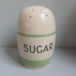 Może przedstawiać: Kremowy ceramiczny cukiernica o zaokrąglonym kształcie. Słowo "SUGAR" jest wydrukowane na czarno, z zielonymi paskami otaczającymi podstawę i paskiem w pobliżu góry. Na górze znajduje się kilka małych otworów.