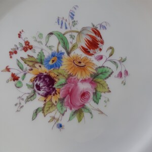 May include: A white ceramic plate with a gold rim features a hand-painted floral design. The bouquet includes a pink rose, a yellow sunflower, a red and orange tulip, and blue and purple wildflowers.