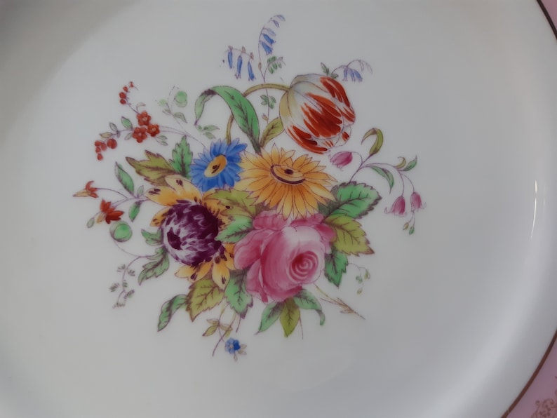 May include: A white ceramic plate with a gold rim features a hand-painted floral design. The bouquet includes a pink rose, a yellow sunflower, a red and orange tulip, and blue and purple wildflowers.