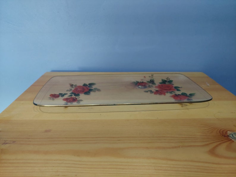 Pode incluir: Uma bandeja de servir de vidro transparente com uma borda dourada e um design floral com rosas vermelhas e folhas verdes.