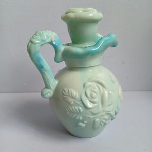 Może przedstawiać: Jasnozielony ceramiczny dzbanek z g&oacute;rą i uchwytem w kształcie r&oacute;ży. Dzbanek posiada wytłaczane wzory r&oacute;ż i jasnoniebieski akcent na uchwycie i g&oacute;rze. Całość jest bogato zdobiona i dekoracyjna.
