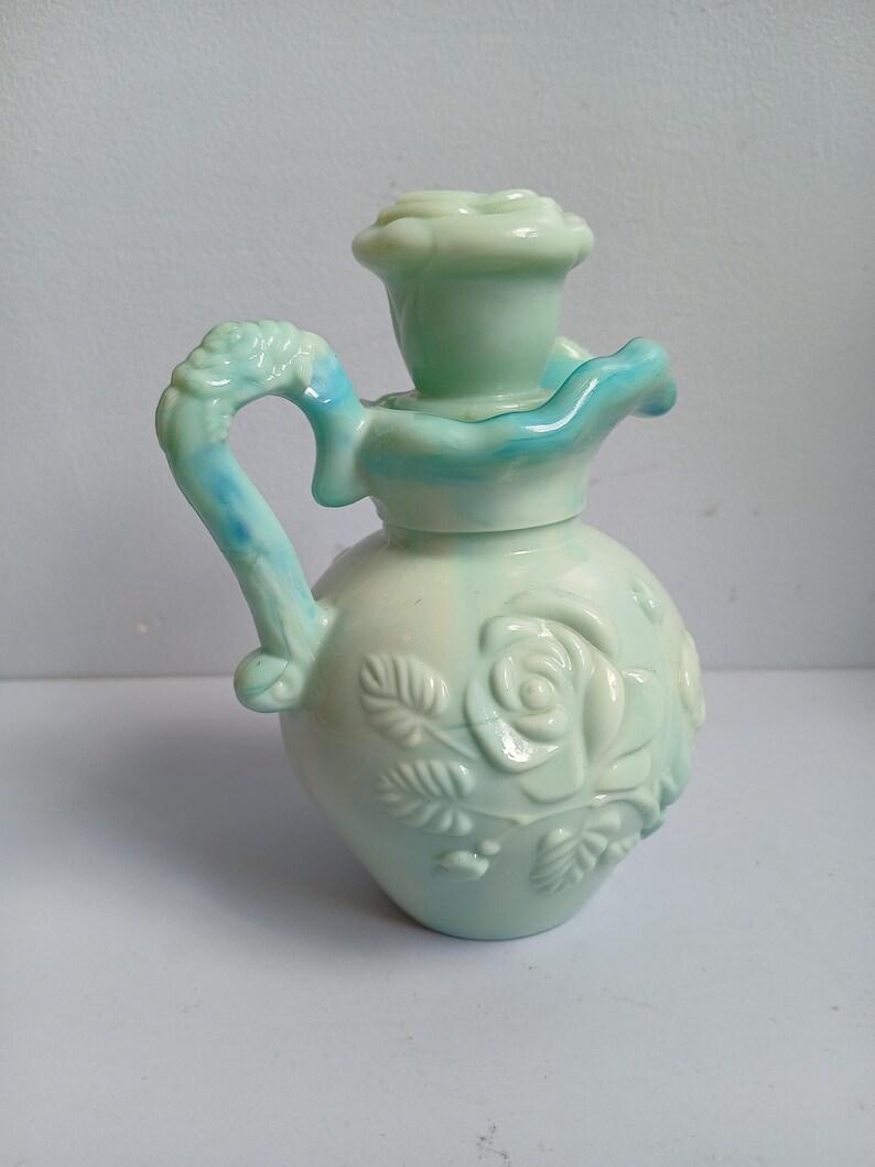 Może przedstawiać: Jasnozielony ceramiczny dzbanek z g&oacute;rą i uchwytem w kształcie r&oacute;ży. Dzbanek posiada wytłaczane wzory r&oacute;ż i jasnoniebieski akcent na uchwycie i g&oacute;rze. Całość jest bogato zdobiona i dekoracyjna.