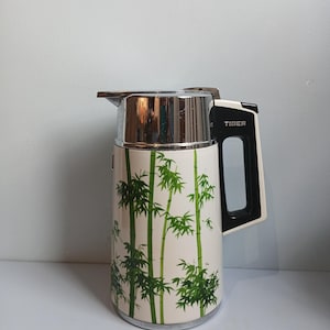 Puede incluir: Un termo aislado vintage con un cuerpo color crema con un diseño de bambú verde. La parte superior y la boquilla son cromadas, y el asa es negra y blanca. La palabra "TIGER" está impresa en el lateral.
