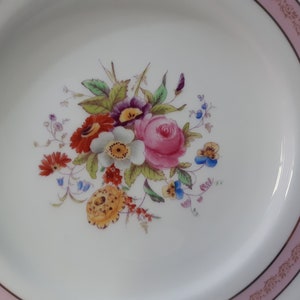May include: A white ceramic plate with a pink and gold rim. The center of the plate features a floral design with pink, red, yellow, blue, and white flowers.