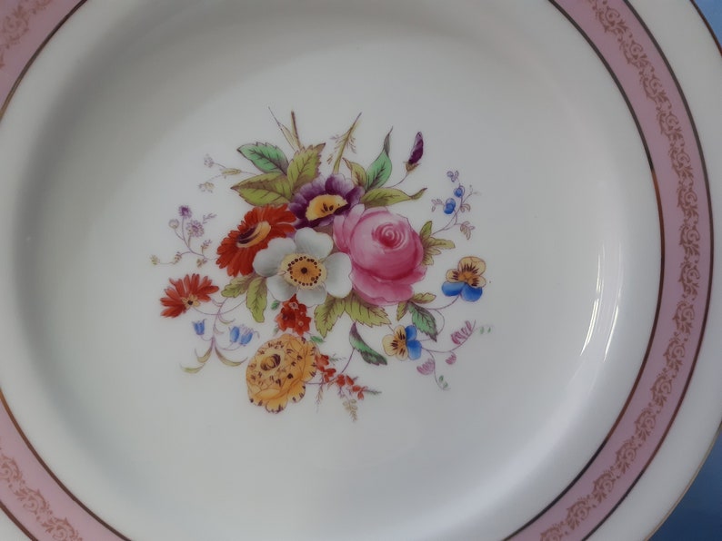 May include: A white ceramic plate with a pink and gold rim. The center of the plate features a floral design with pink, red, yellow, blue, and white flowers.
