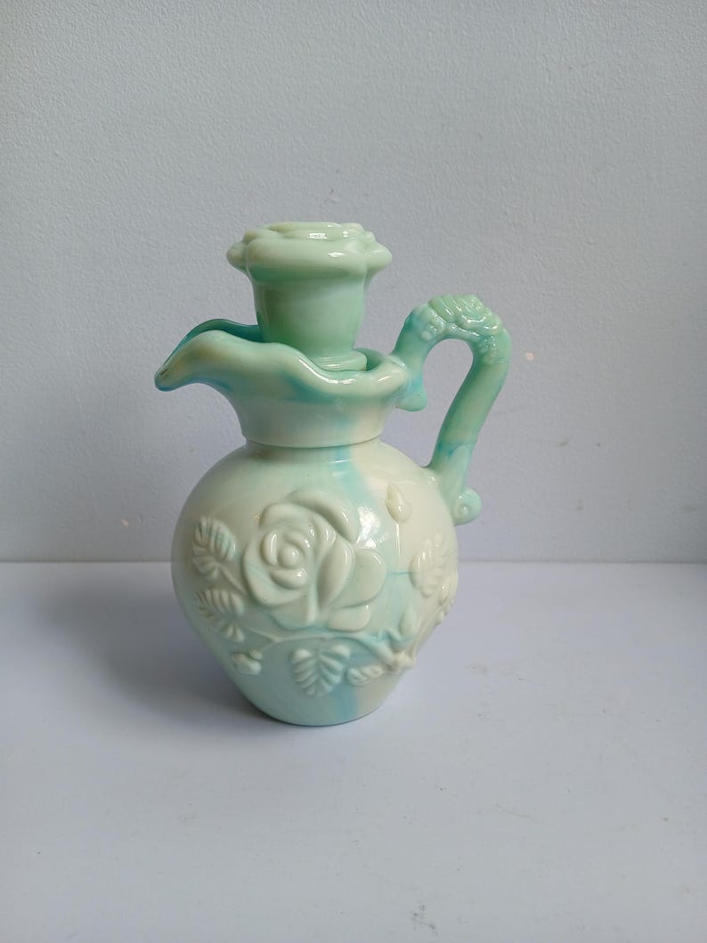 Może przedstawiać: Ceramiczny dzban z kwiatowym wzorem i uchwytem. Dzban jest pomalowany w jasnej zieleni i bieli, z reliefem r&oacute;ży z przodu. Uchwyt ma dekoracyjny, teksturowany wz&oacute;r.