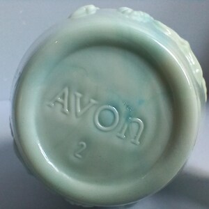 Może przedstawiać: Widok od spodu jasnoniebieskiej ceramicznej butelki z wytłoczonym napisem "AVON" i cyfrą "2". Butelka ma zaokrąglony kształt i lekko teksturowaną powierzchnię. Ceramika ma błyszczące wykończenie.