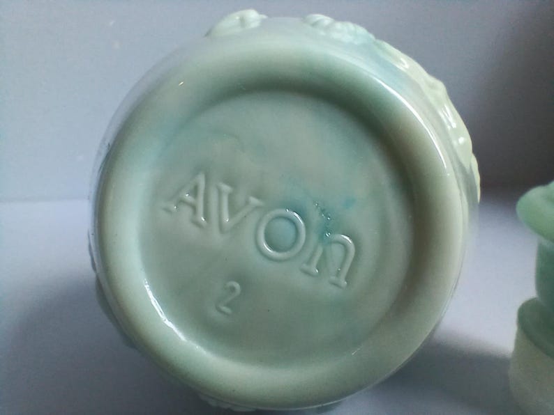 Może przedstawiać: Widok od spodu jasnoniebieskiej ceramicznej butelki z wytłoczonym napisem "AVON" i cyfrą "2". Butelka ma zaokrąglony kształt i lekko teksturowaną powierzchnię. Ceramika ma błyszczące wykończenie.