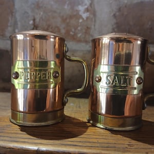 Puede incluir: Un par de saleros y pimenteros de cobre con detalles de latón. Los saleros y pimenteros tienen un diseño similar a una taza con asas y etiquetas de latón que dicen "PEPPER" y "SALT". El cobre tiene un acabado pulido y los saleros y pimenteros están sobre una superficie de madera.