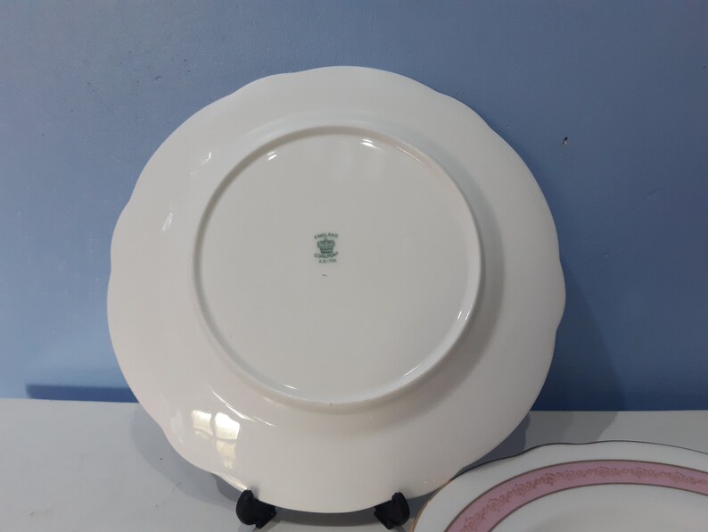 May include: White ceramic dinner plate with a scalloped edge and a green and white logo that reads "Landau Fine China England" with a crown above the text.