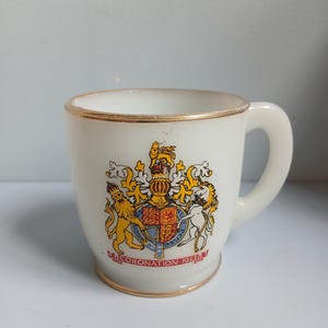 Op de afbeelding: Witte keramische mok met een gouden rand en oor. De mok heeft een kleurrijk embleem met een kroon, leeuwen en de tekst "CORONATION 1953". Een vintage verzamelobject.
