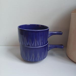 Puede incluir: Dos cuencos de sopa de cerámica azul cobalto apilados con asas. Cada cuenco presenta un patrón vertical texturizado. Los cuencos están sobre una superficie blanca, con un fondo neutro y un vistazo a un recipiente de cerámica beige.