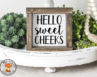 May include: A white wooden sign with the text "HELLO sweet CHEEKS" in black cursive lettering. The sign is framed in a dark brown wood frame and is sitting on a white tray with green plants and a wooden bead garland.