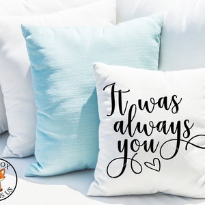 Pode incluir: Travesseiro branco com texto em itálico preto que diz "It was always you" com um pequeno símbolo de coração.