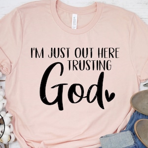 May include: A pink t-shirt with the text "I'm just out here trusting God" in black lettering with a small heart.