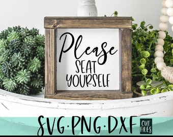 May include: A white wooden sign with black text that reads "Please seat yourself". The sign is sitting on a white surface with green plants on either side.