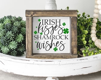 May include: A wooden sign with the text "Irish kisses shamrock wishes" in black script with green shamrock accents. The sign is sitting on a white tray with green plants and white beads.