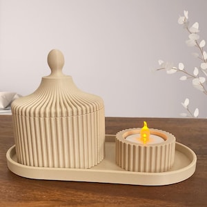 May include: A beige decorative set comprising a lidded jar and a candle holder with a flickering LED candle, both with vertical ribbed detailing. The set is on an oval tray, creating a cohesive aesthetic for home decor.