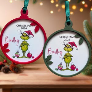 May include: Two round Christmas ornaments with green ribbons. One has a red frame, the other a dark green frame. Both feature a green character in a Santa hat, the text 'CHRISTMAS 2026', the name 'Bradley' in red script, and small holly leaf accents.