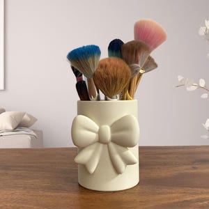 May include: A cream-coloured cylindrical makeup brush holder with a 3D bow design. The holder contains various makeup brushes with colourful bristles, including blue, pink, and brown. The holder is on a wooden surface.