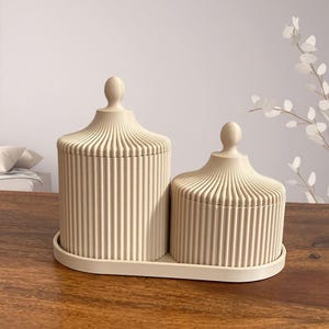 May include: Two beige decorative jars with lids, one large and one small, sit on a matching tray. The jars have a ribbed design and a rounded top. The set is displayed on a wooden surface.