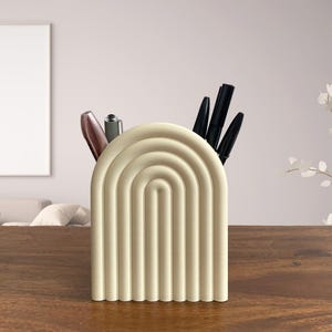 May include: A cream-coloured, arched desk organiser featuring a series of concentric semi-circles. The organiser holds several pens and pencils. The desk organiser is made of a solid material and sits on a wooden surface.