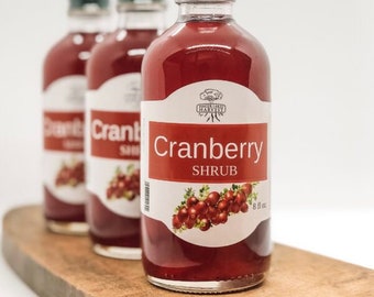 Op de afbeelding: Drie glazen flessen cranberry-siroop, elk met een wit etiket met een rode cranberry-illustratie en de tekst "Cranberry Shrub".