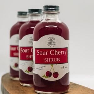 May include: Three glass bottles of sour cherry shrub, a red, vinegar-based syrup, with a white label that reads "Sour Cherry Shrub" and features a red cherry illustration.