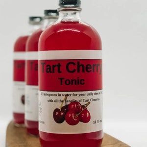 May include: A glass bottle of red tart cherry tonic with a white label that reads "Tart Cherry Tonic" and "2 Tablespoons in water for your daily dose of antioxidants with all the benefits of Tart Cherries". The bottle is sitting on a wooden surface.