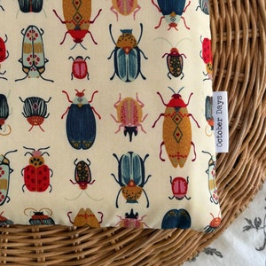 May include: A cream-coloured pillow with a repeating pattern of colourful beetles. The pillow has a white tag with the text "October Days" on it.