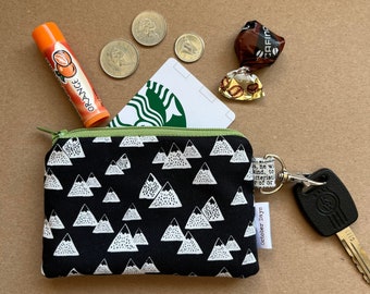 May include: A black and white zippered pouch with a mountain pattern. The pouch has a green zipper and a small tag that says "October Days". The pouch is lying on a brown surface with a Starbucks gift card, a lip balm, and some coins.