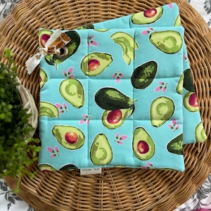 Può includere: Un paio di presine imbottite blu con un motivo di avocado su sfondo azzurro chiaro. Le presine hanno un'etichetta bianca con il testo "October Days" su di esse.