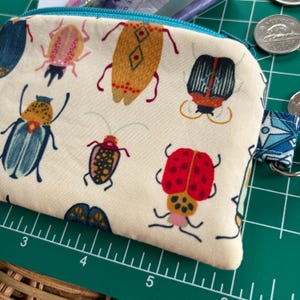 May include: A white fabric coin purse with a teal zipper and a colourful bug pattern. The bugs are red, yellow, blue, and green. The purse has a small loop on the side.