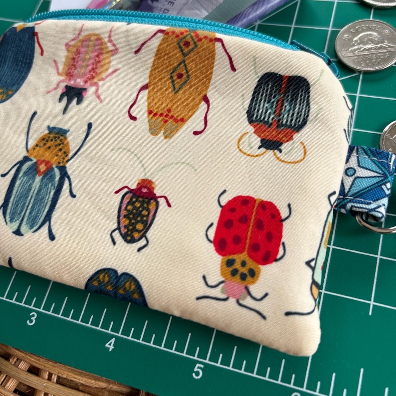May include: A white fabric coin purse with a teal zipper and a colourful bug pattern. The bugs are red, yellow, blue, and green. The purse has a small loop on the side.