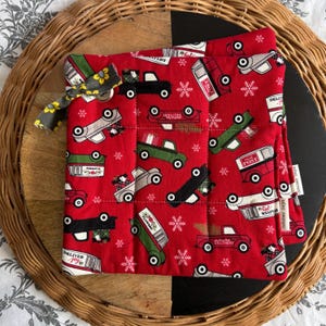 Peut inclure: Manique matelassée rouge avec un motif répété de camions vintage transportant des cadeaux et des flocons de neige. La manique a une boucle en tissu et une étiquette "October Days".