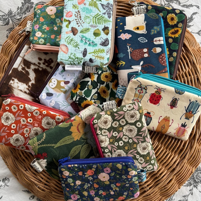May include: A collection of small zippered pouches with various floral, animal, and insect patterns. The pouches are made of fabric and have a variety of colours, including green, blue, red, and brown. The pouches are arranged in a wicker basket.
