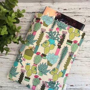 May include: A cream-coloured fabric book cover with a repeating pattern of green, pink and blue cacti in pots. The cover has a button closure and is designed to fit a standard-sized book.