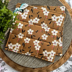 May include: Two brown quilted pot holders with a white floral pattern. The pot holders have a white trim and a small loop on the top.