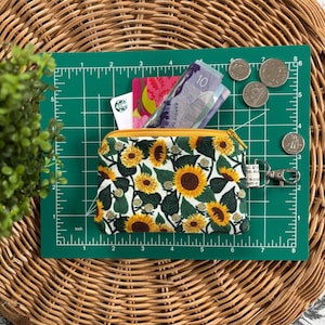 Peut inclure: Une petite pochette zippée avec un imprimé tournesol. La pochette est en tissu et possède une fermeture éclair jaune. La pochette est ouverte et contient de l'argent liquide, des cartes-cadeaux et des pièces de monnaie. La pochette est posée sur un tapis de coupe vert avec une règle.