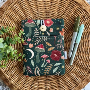 May include: A dark green fabric pouch featuring a mushroom and floral design, secured with a button and loop. Two pens, one grey and one light green, are placed beside the pouch on a woven surface. The pouch is decorated with a whimsical pattern.