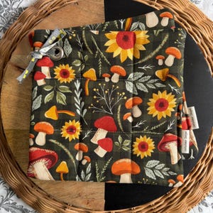 Op de afbeelding: Een gewatteerde boekenhoes met een patroon van zonnebloemen en paddenstoelen in rood, oranje, geel en groen. De hoes heeft een grijs lint en een metalen oogje. De woorden "October Days" staan op een klein label.