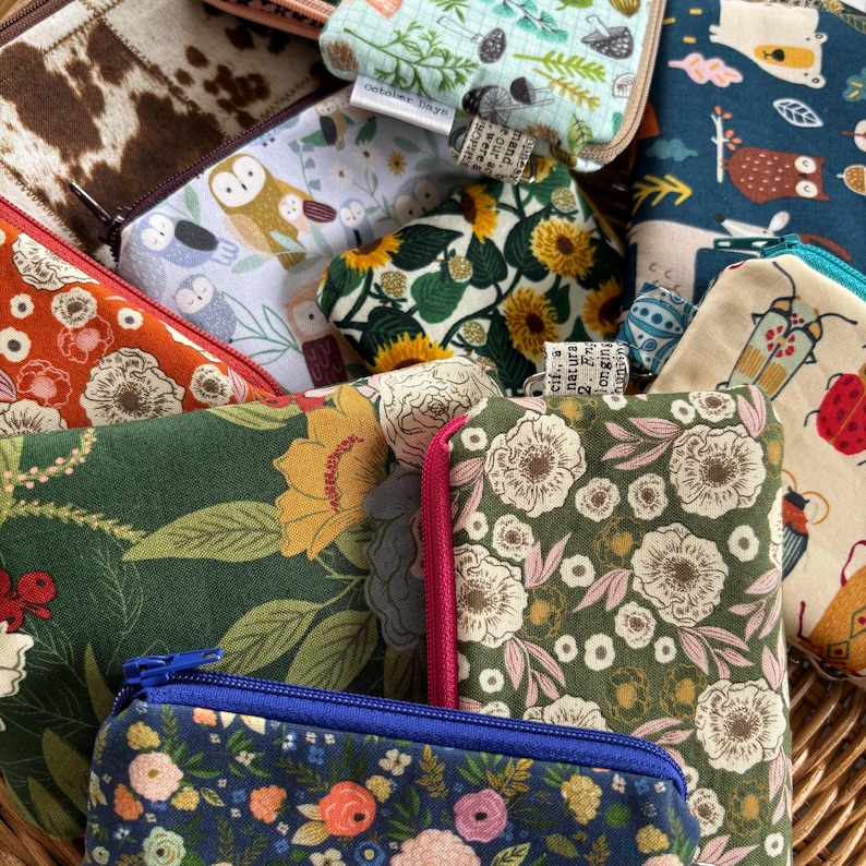 May include: A collection of small fabric pouches with various floral and nature-themed prints. The pouches are made of cotton fabric and have zipper closures. The pouches are arranged in a pile, with some of the pouches overlapping each other.
