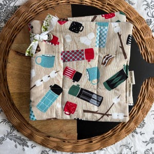 May include: A beige quilted pot holder with a camping theme. It features illustrations of mugs, marshmallows, and thermoses in red, black, and teal. The pot holder is tied with a white and green patterned ribbon.