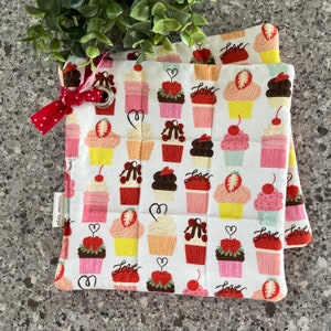 May include: Two white quilted pot holders with a red and white gingham ribbon. The pot holders feature a repeating pattern of cupcakes with cherries, strawberries, and chocolate frosting. The words "Love" are printed on the pot holders.