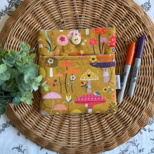 May include: Mustard fabric pouch featuring a mushroom design. The pouch has a flap secured by a wooden button. Two Sharpie markers, orange and purple, are placed beside the pouch. The pouch rests on a woven wicker surface.