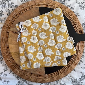 May include: Mustard yellow quilted pot holders with a white rose pattern. The pot holders are folded on a wooden serving board with a black handle, set on a woven tray. A fabric tie with a metal grommet is attached to the pot holders.
