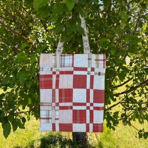 May include: A rectangular tote bag constructed from a patchwork of fabric squares featuring a red, white, and light green plaid pattern alongside white panels with a subtle circular line design, complete with two matching fabric shoulder straps.